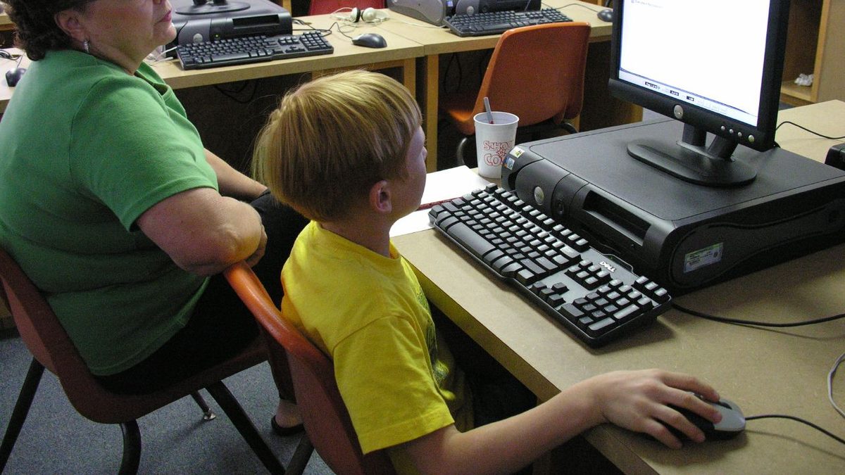 A child's hand on a mouse in a school computer lab, a grown-up sitting alongside.