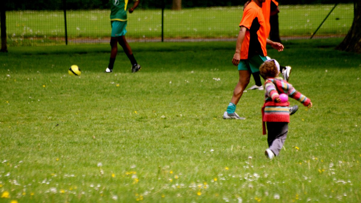 A kickabout in a park: players in bibs, a ball in the grass, a toddler running after them.
